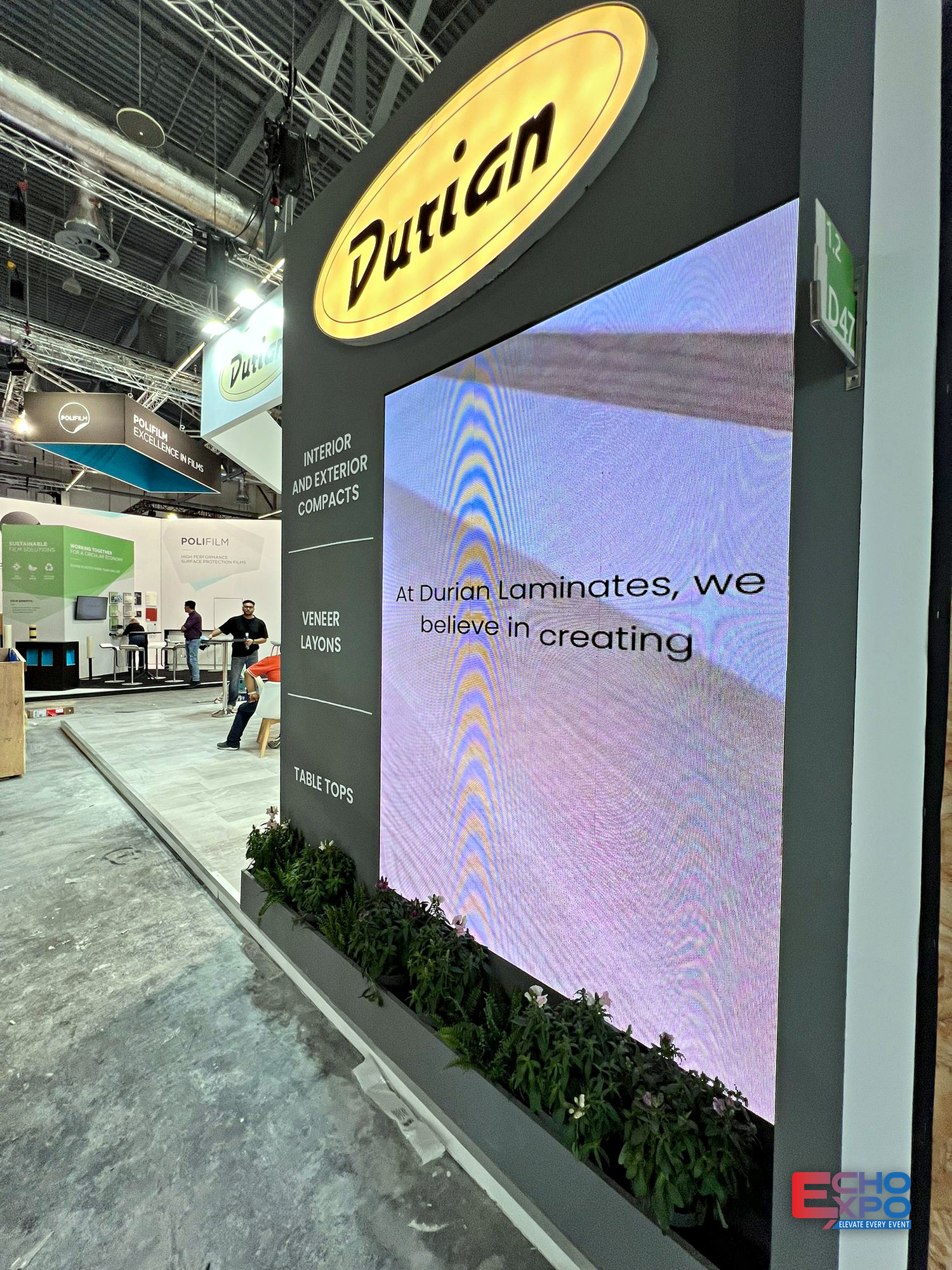 Large LED video wall on the Durian Laminates stand at interzum Cologne 2025, set into a dark wall above a planted base.
