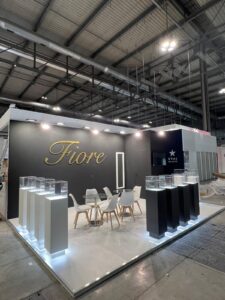 Fiore eyewear stand at MIDO, Fiera Milano — black feature wall with gold three-dimensional lettering, illuminated glass display plinths and a white lit platform floor