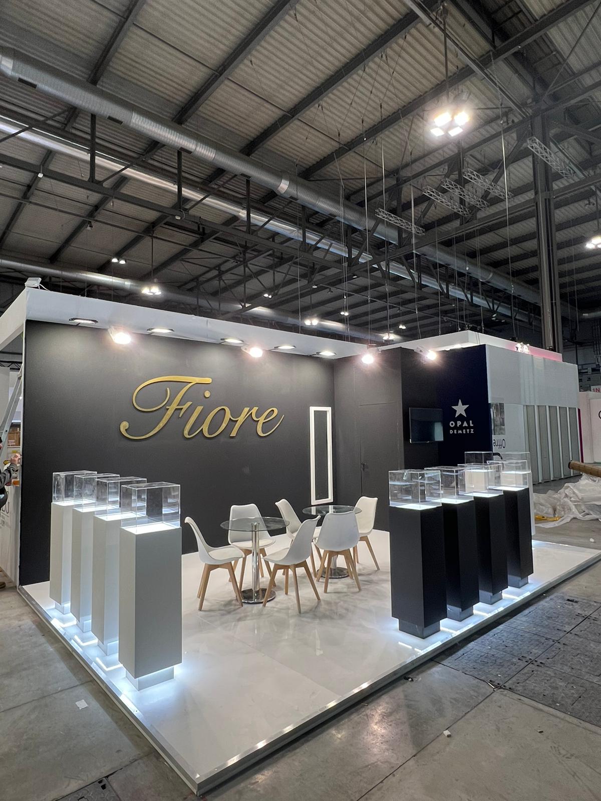 Fiore eyewear stand at MIDO, Fiera Milano — black feature wall with gold three-dimensional lettering, illuminated glass display plinths and a white lit platform floor