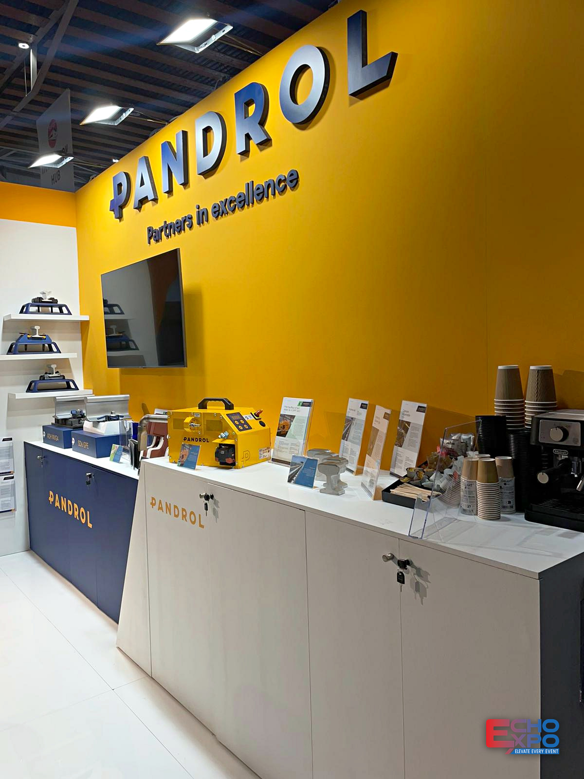 Yellow PANDROL wall with dimensional cut lettering, a wall-mounted screen and rail fastening products displayed on shelves above lockable counters.