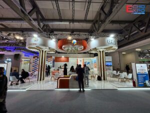 Finished SP Accure Labs stand at CPHI Milan 2026 from the aisle, with two tree columns, a suspended orange ring and a reception desk.