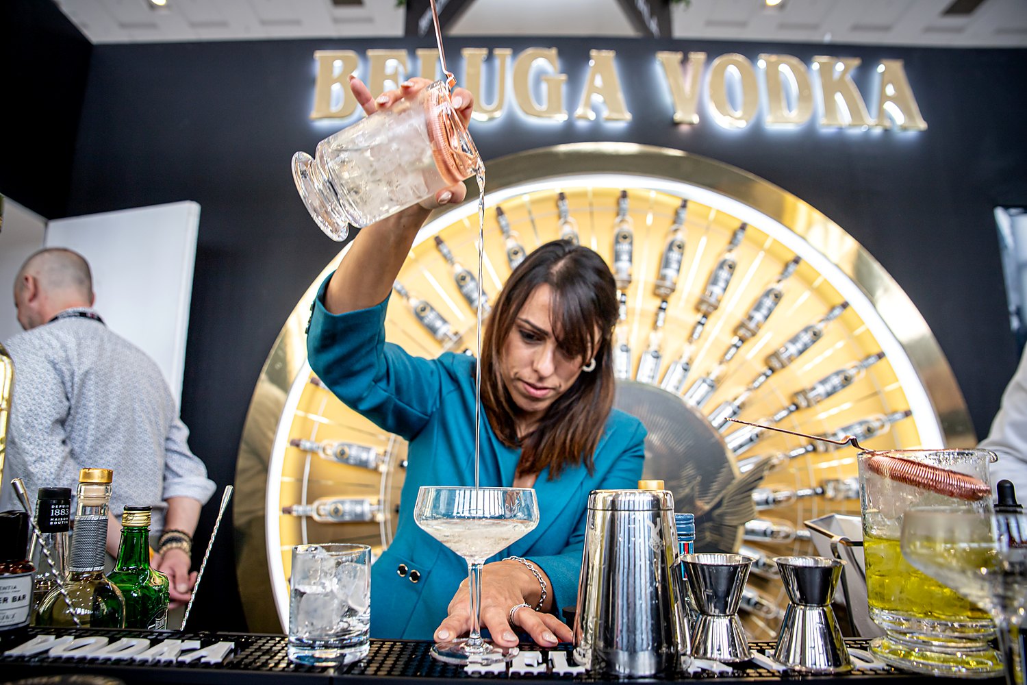 A bartender pouring a cocktail at the Beluga Noble Vodka bar at Bar Convent Berlin 2023, in front of the gold sunburst bottle wall.