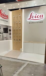 Leica Microsystems stand at the ESCRS 2022 congress in Milan: white walls with backlit red Leica logos, a timber feature panel and a white storage counter.