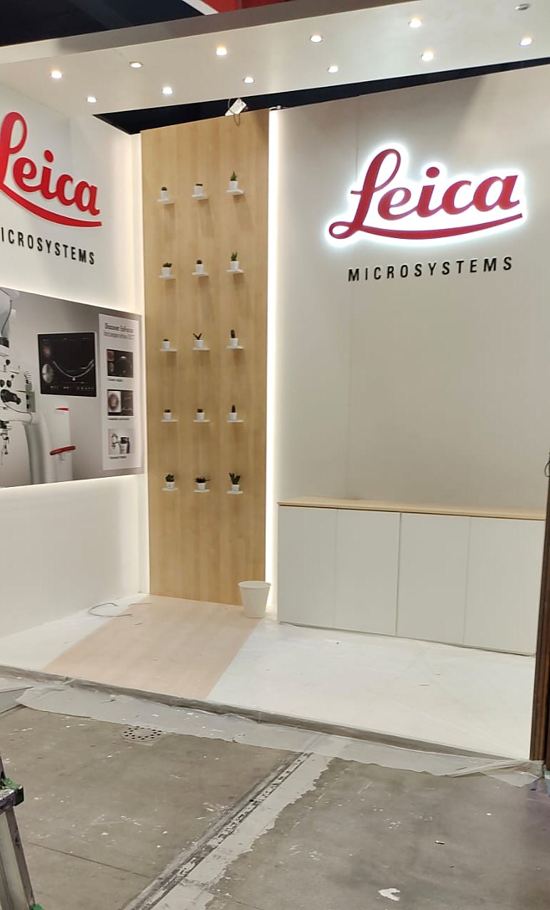 Leica Microsystems stand at the ESCRS 2022 congress in Milan: white walls with backlit red Leica logos, a timber feature panel and a white storage counter.