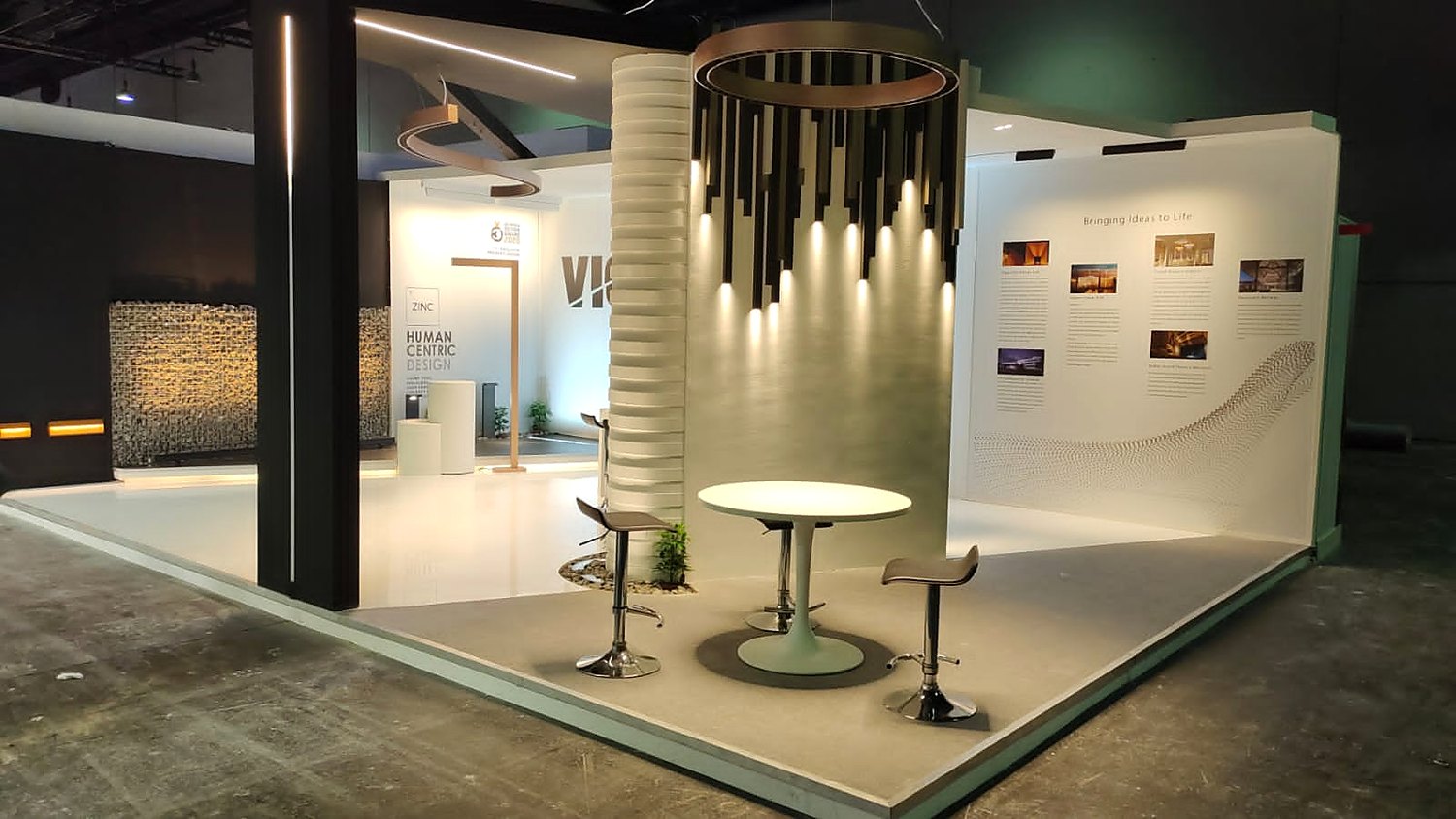 Inside the VICE stand at Light + Building 2022: a cluster of slim pendant lights hanging from a ring above a round illuminated table, with a ribbed white column and framed images on the wall.