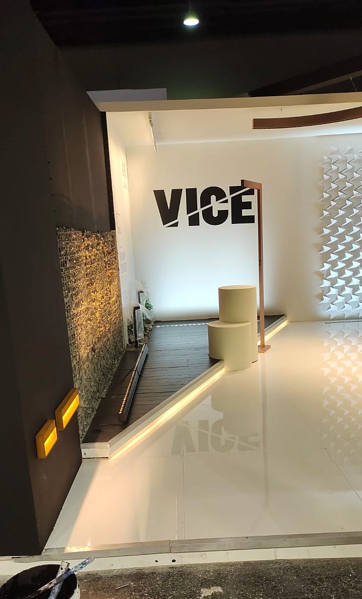 Corner view of the VICE stand at Light + Building Frankfurt, with the black VICE logo on a white wall, a warm-lit stone gabion wall and a light line along the gloss floor.