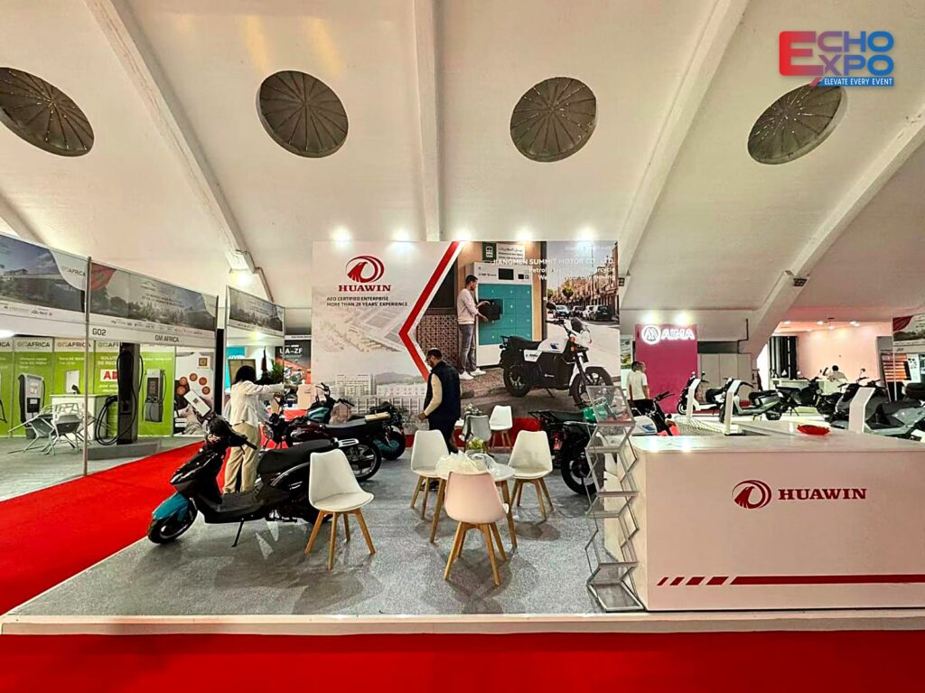 HUAWIN (Jiangmen Summit Motor) exhibition stand H07–H08 on show day at EVS Mobility Expo Morocco 2026, OFEC Casablanca: 36 m² with a red and white graphic back wall, white HUAWIN counter, motorcycles and chairs on a grey floor beside the red-carpet aisle, built by Echo Expo.