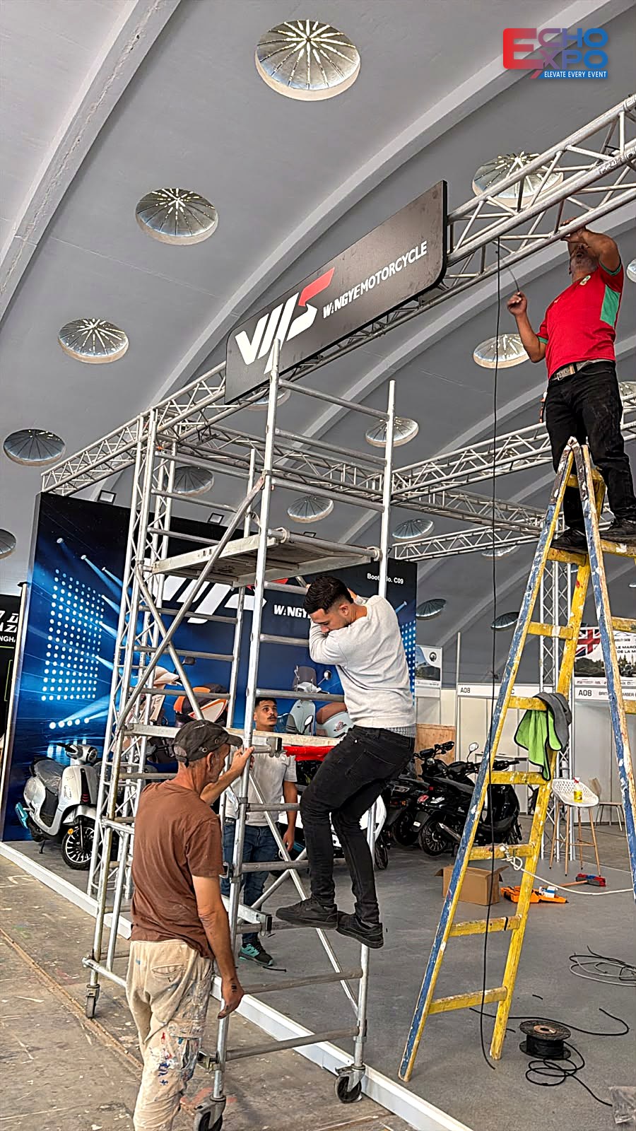 Crew on a mobile scaffold fixing the suspended WANGYE fascia banner to the truss frame, EVS Mobility Expo Morocco 2026 build-up.