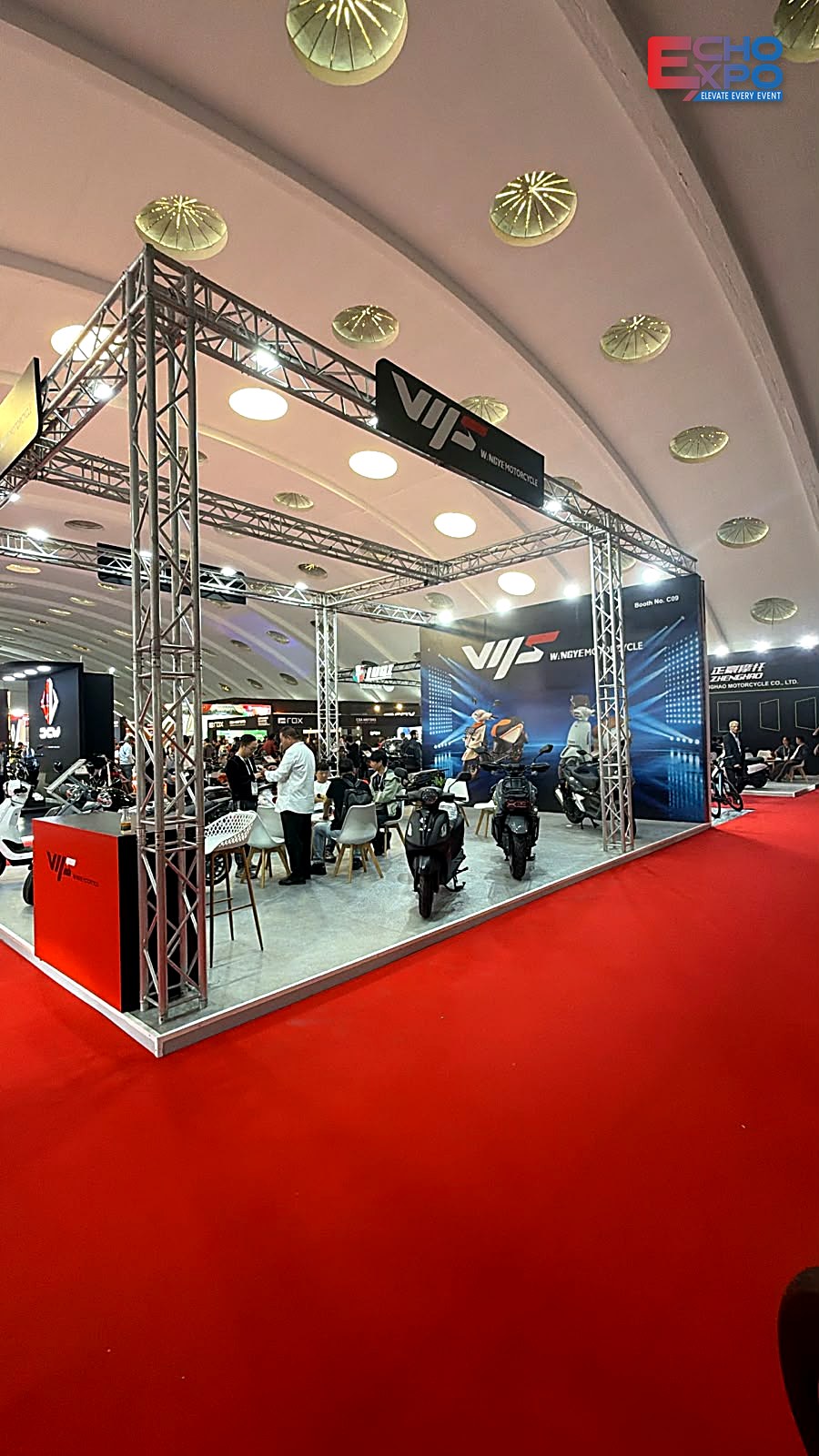 Corner view of the WANGYE Motorcycle stand at EVS Mobility Expo Morocco 2026: truss frame with LED lights, scooters on a grey floor and the red reception counter.