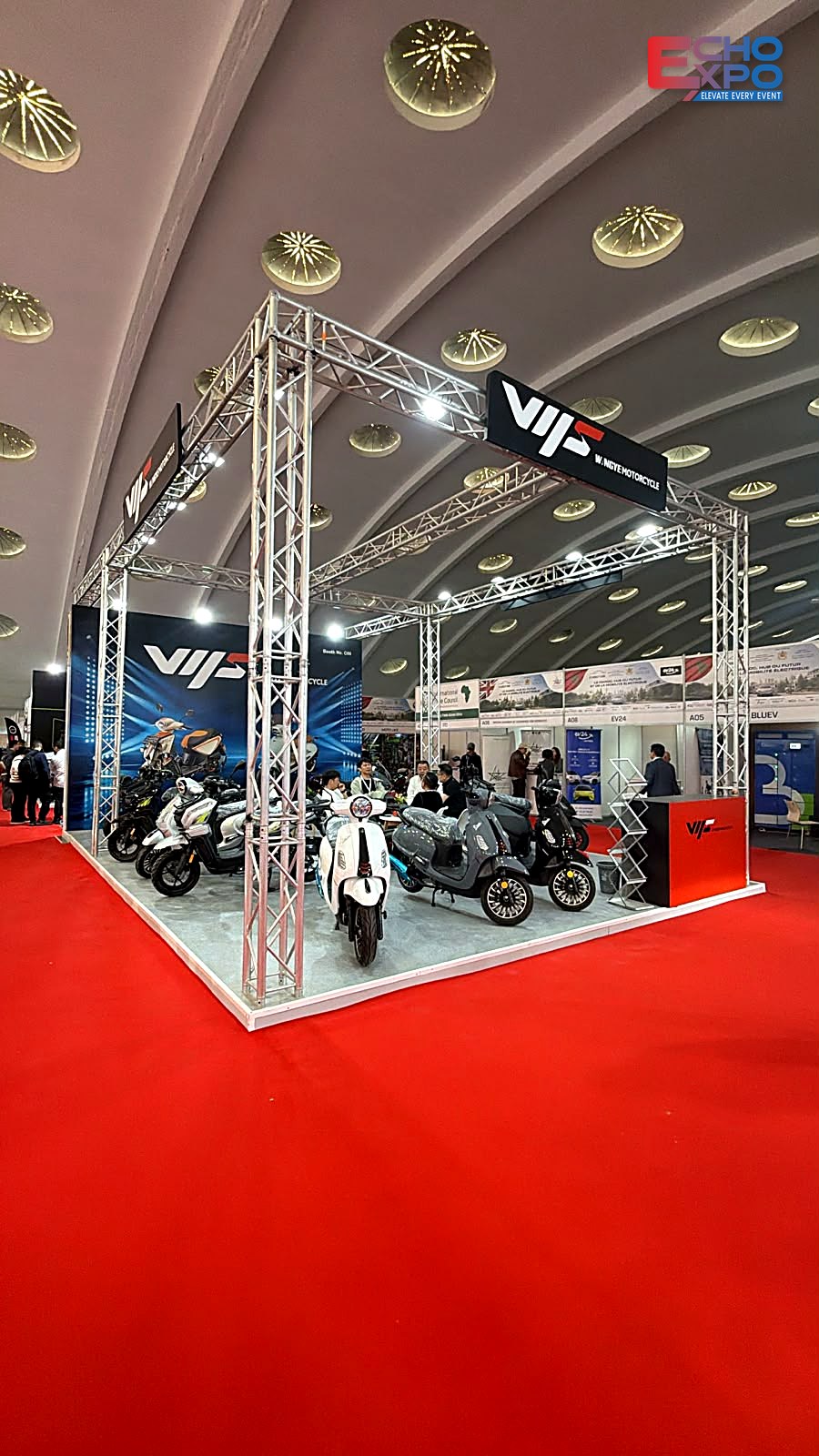 WANGYE Motorcycle exhibition stand C09 at EVS Mobility Expo Morocco 2026, OFEC Casablanca: 54 m² open stand under a lit 30 cm truss frame with suspended fascia, blue fabric back wall and red counter, by Echo Expo.