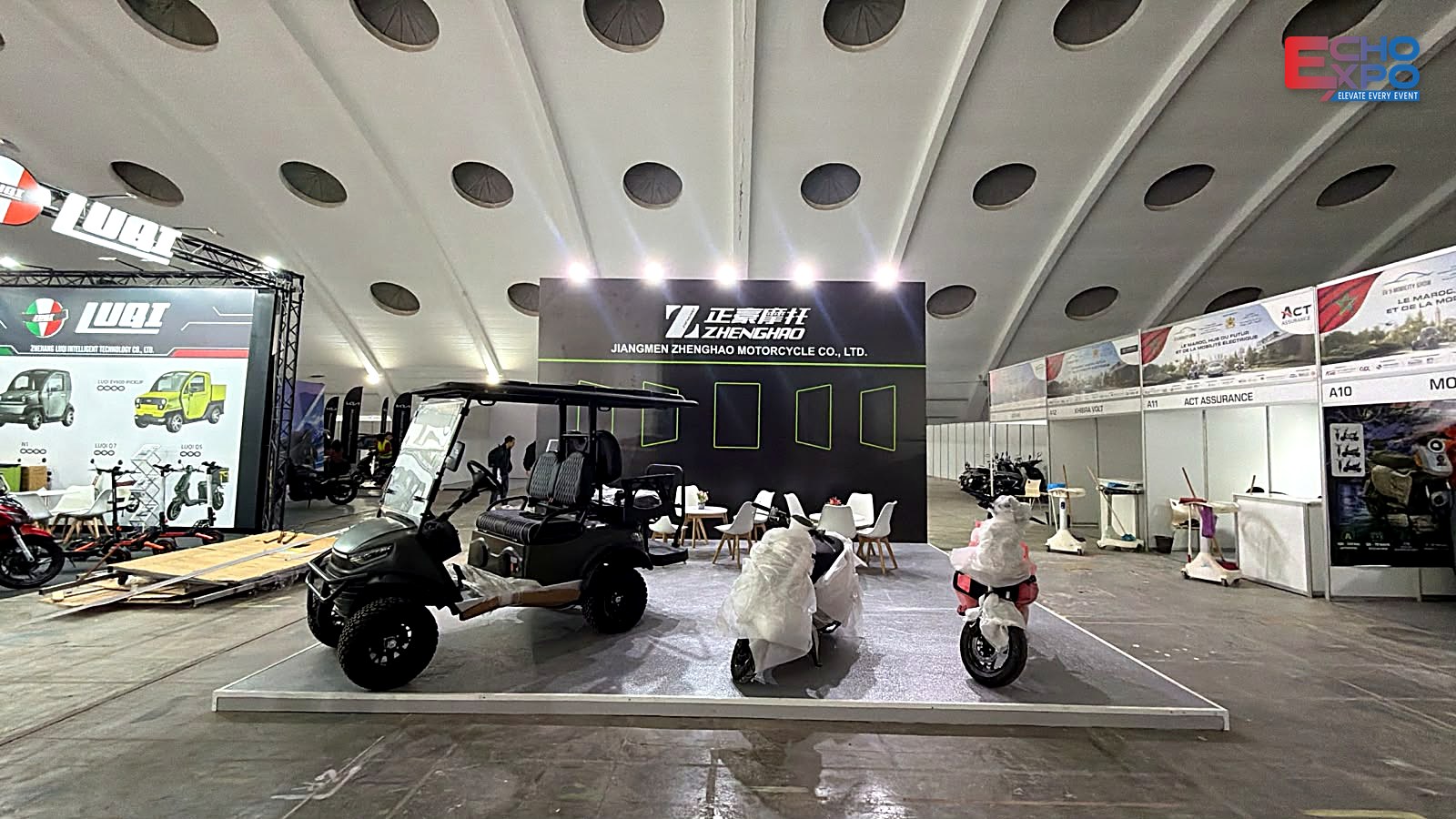 ZHENGHAO stand finished and handed over: exhibitor vehicles arriving on the raised floor the day after build-up, EVS Mobility Expo Morocco 2026.