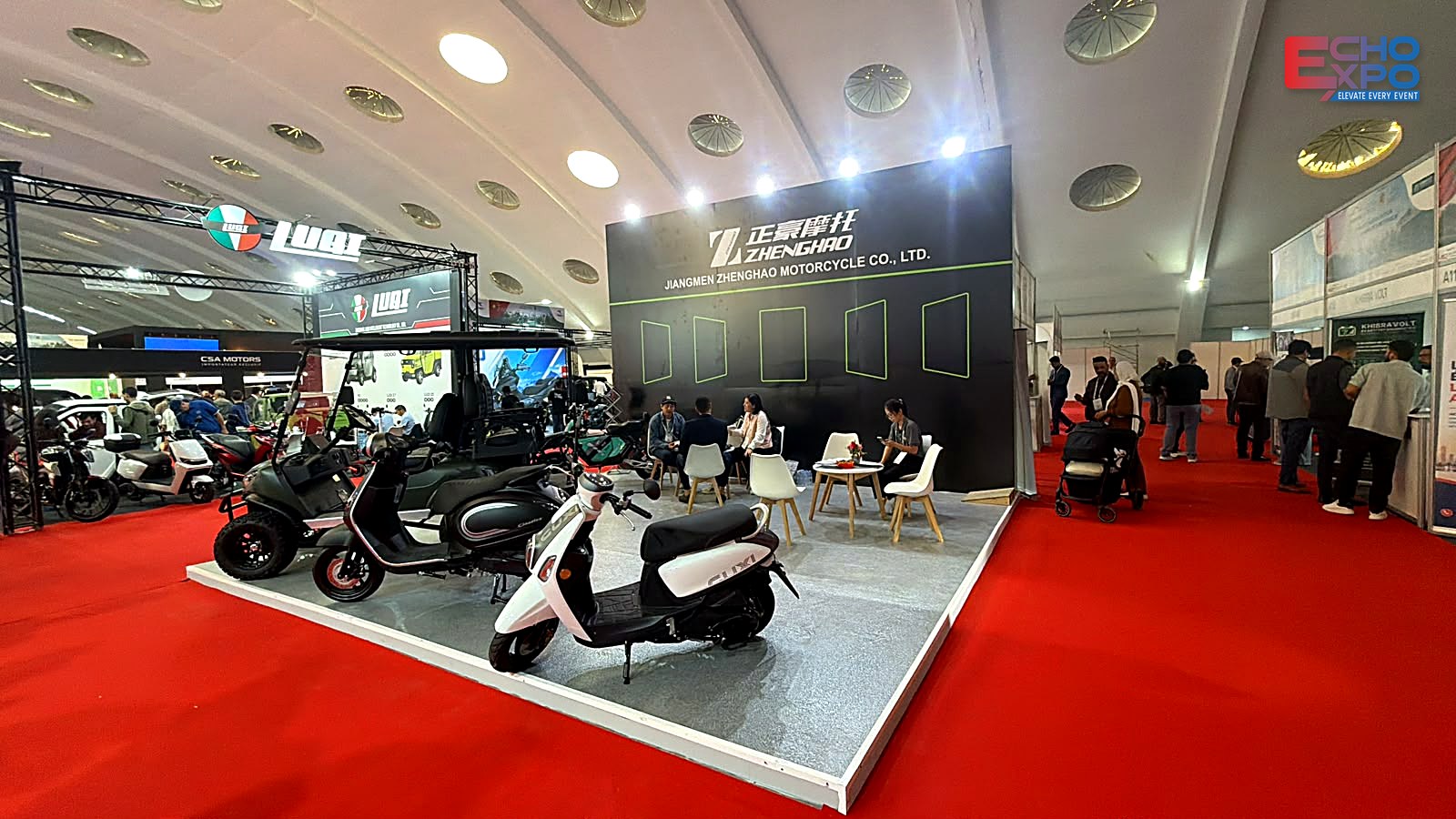 ZHENGHAO stand at EVS Mobility Expo Morocco 2026 during the show: visitors in the aisle, scooters at the front edge and a meeting area by the black back wall.